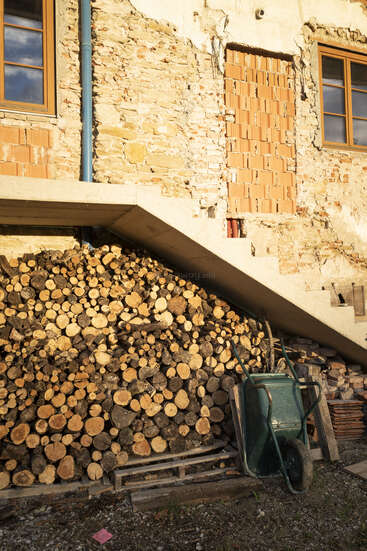 Una gran pila de leña se apila ordenadamente bajo las escaleras exteriores, junto a una vieja casa de ladrillo desgastado. Una carretilla verde se apoya en la pila de leña.