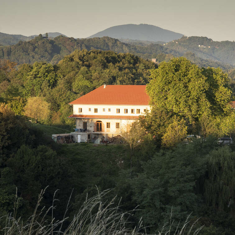 Una gran casa con tejado rojo se alza rodeada de frondosos árboles verdes y colinas, bañada por la cálida luz del sol, con las montañas al fondo.