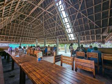 Cette image montre des personnes assises sur des chaises en bois sous une grande structure aérée au toit de chaume. Les tables sont en bois et l'atmosphère est ouverte et détendue.