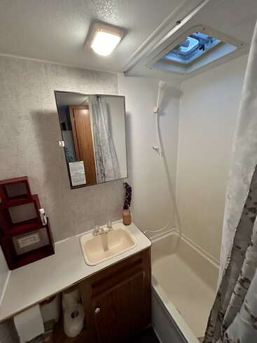 Esta imagen muestra un cuarto de baño compacto de una autocaravana con un lavabo pequeño, un armario con espejo, una ducha con cabezal de mano, una claraboya, un mueble de baño, un dispensador de jabón y estantes de pared.