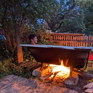 The image depicts a man sitting in a clawfoot bathtub outdoors, surrounded by a fire pit and wooden planks, with a fence and trees in the background.