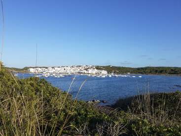 Das Bild zeigt eine ruhige Küstenszene mit einer Ansammlung weißer Gebäude auf einem Hügel, die ein ruhiges Gewässer mit Booten und üppiger Vegetation im Vordergrund überblicken.