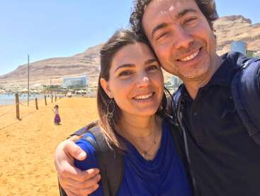 Das Bild zeigt einen Mann und eine Frau, die für ein Selfie an einem Strand vor einem bergigen Hintergrund posieren. Beide tragen Rucksäcke und lächeln in die Kamera.