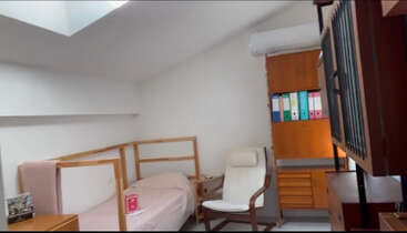 Das Bild zeigt ein Zimmer mit einem Bett, einem Schreibtisch und einem Stuhl, mit einem Fenster und einer Tür, mit einem Bücherregal und einem Tisch und einer weißen Wand im Hintergrund.