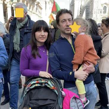 A family stands together in a crowded outdoor setting. The man holds a child, while the woman stands beside a stroller. Faces are partially obscured for privacy.