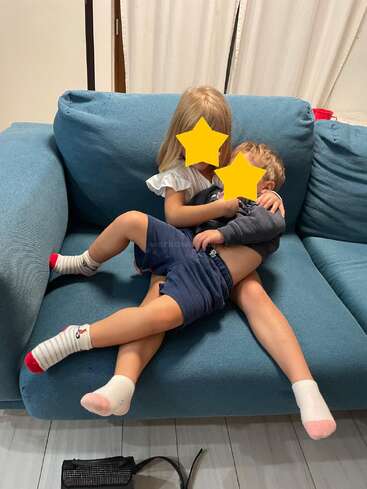 Zwei kleine Kinder sitzen zusammen auf einem blauen Sofa und kuscheln. Ihre Gesichter sind mit Sternchenaufklebern bedeckt. Sie wirken entspannt und bequem und tragen legere Kleidung.