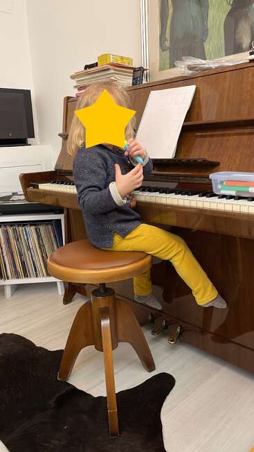 Ein kleines Kind sitzt auf einem Holzschemel an einem Klavier, umgeben von Büchern, Schallplatten und Kunst. Das Kind hält einen blauen Marker in der Hand.