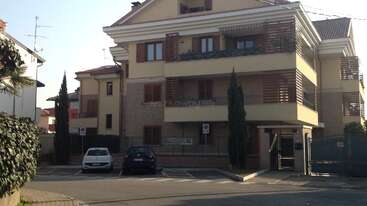 Ein modernes dreistöckiges Wohnhaus mit Balkonen und Topfpflanzen. Vor dem Haus sind zwei Autos geparkt, es hat eine Toreinfahrt und hohe, schlanke Bäume säumen die Fassade.