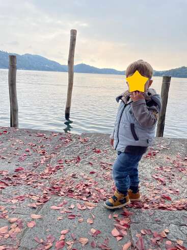 Ein kleines Kind steht auf einem mit Laub bedeckten Steinweg an einem friedlichen See, umgeben von Holzpfählen und Bergen im Hintergrund unter einem bewölkten Himmel.