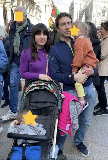 A family stands together in a crowded outdoor setting. The man holds a child, while the woman stands beside a stroller. Faces are partially obscured for privacy.