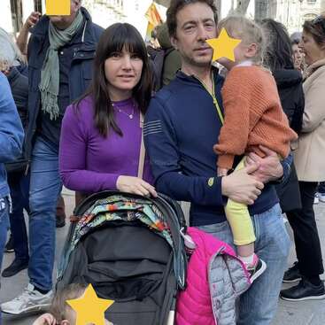 Eine Familie steht zusammen in einem überfüllten Außenbereich. Der Mann hält ein Kind, während die Frau neben einem Kinderwagen steht. Die Gesichter sind teilweise unkenntlich gemacht, um die Privatsphäre zu wahren.