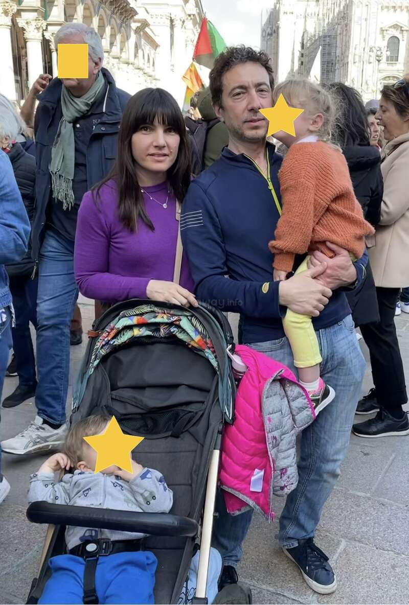 Eine Familie steht zusammen in einem überfüllten Außenbereich. Der Mann hält ein Kind, während die Frau neben einem Kinderwagen steht. Die Gesichter sind teilweise unkenntlich gemacht, um die Privatsphäre zu wahren.