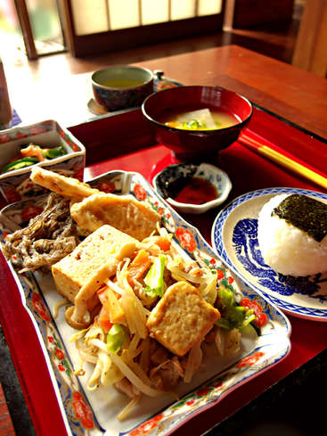 A imagem retrata uma refeição japonesa vibrante em uma bandeja vermelha, com uma variedade de pratos, incluindo sopa de missô, arroz e legumes fritos, dispostos em uma mesa de madeira.
