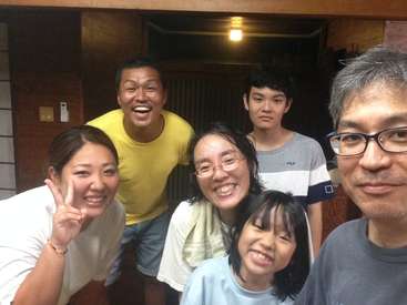 A imagem mostra uma família de seis pessoas posando para uma selfie, sorrindo e olhando para a câmera, com uma porta de madeira atrás deles.