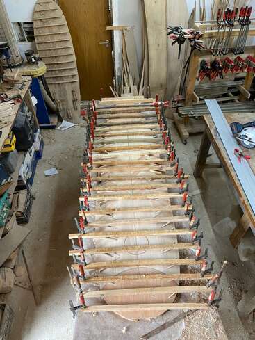 L'image représente un atelier encombré avec, au centre, une structure en bois comportant des pinces et des outils, entourée de matériaux et d'équipements divers.