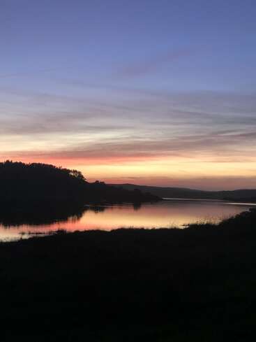 L'image représente un coucher de soleil serein sur une étendue d'eau tranquille, avec une silhouette d'arbres et de collines en arrière-plan, évoquant un sentiment de paix.