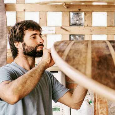 L'image représente un homme aux cheveux et à la barbe foncés, vêtu d'un t-shirt gris, debout devant un mur en bois, tenant un objet en bois de la main droite.