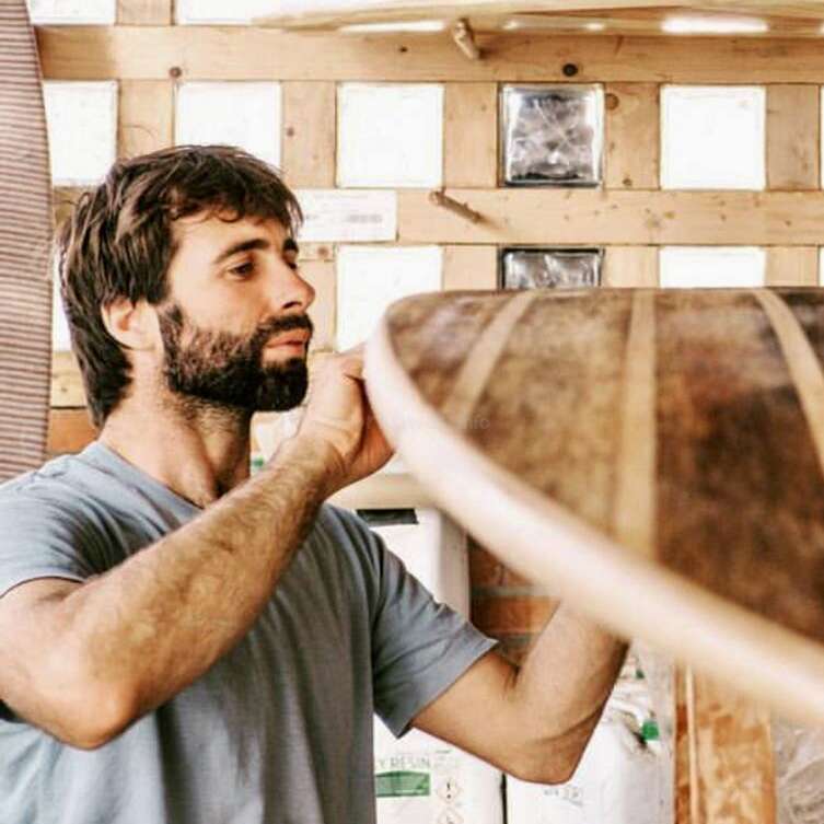 L'image représente un homme aux cheveux et à la barbe foncés, vêtu d'un t-shirt gris, debout devant un mur en bois, tenant un objet en bois de la main droite.