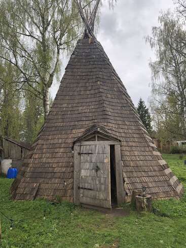 This image shows a large, conical wooden hut with a shingled roof, surrounded by green grass and trees under a cloudy sky. Door partially open.