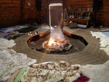 A cozy wooden hut with a central fire pit, surrounded by animal hides and cushions, creating a warm space for gathering, relaxation, and conversation. Natural decor accents.