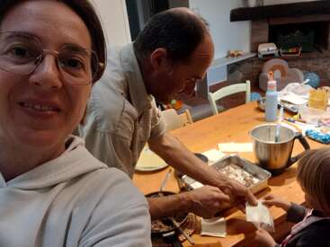 A woman smiles at the camera while a man and a child prepare food together at a wooden table in a cozy, homey kitchen setting.