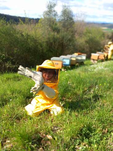 A child wearing a bright yellow beekeeping suit and gloves kneels in the grass, surrounded by green bushes and several colorful beehives in the background.
