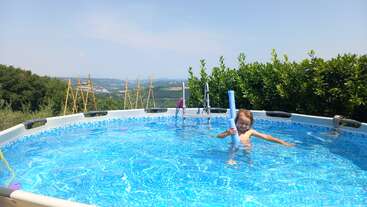 A child is playing with a blue pool noodle in a bright, round outdoor swimming pool surrounded by green bushes, under a clear, sunny sky.