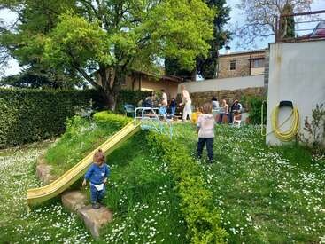 Children play on a slide in a grassy, flower-filled yard while a group of adults gathers at tables in the background, enjoying a sunny day.