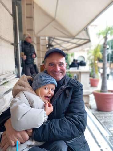 A man and a young girl warmly dressed in winter jackets and hats sit together outside a café, smiling joyfully while enjoying a cozy moment together.
