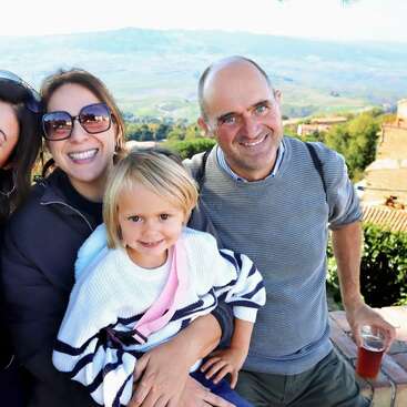 A smiling family enjoys a sunny day outdoors with scenic mountains in the background. The child sits on a woman's lap, while a man holds a drink.