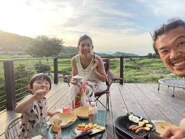 The image depicts a family of three enjoying a meal on a deck, with a serene landscape of greenery and hills in the background under a cloudy sky.