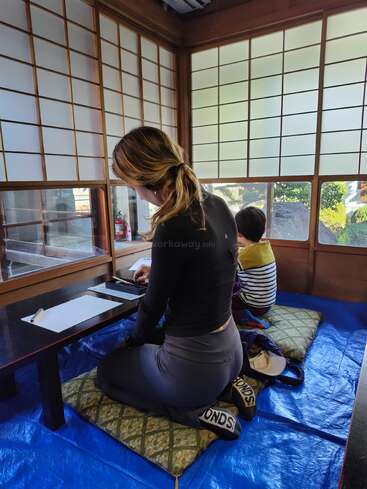 Uma mulher e uma criança estão sentadas em almofadas em uma mesa baixa em uma sala tradicional japonesa com biombos shoji, fazendo uma atividade juntas em um tapete azul.