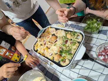 Mehrere Leute versammeln sich um einen Tisch und bereiten Takoyaki in einem elektrischen Takoyaki-Gerät zu. Zutaten wie Frühlingszwiebeln, Tintenfisch und Mais sind zu sehen und sorgen für eine festliche Stimmung.