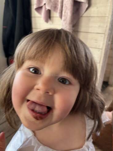 A little girl with brown hair smiles playfully at the camera, sticking out her tongue. She seems joyful and carefree, wearing a white top indoors.