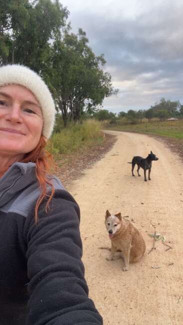 Una mujer sonriente con gorro blanco se toma un selfie en un camino de tierra, acompañada de dos perros, rodeada de árboles bajo un cielo nublado. Un día tranquilo.