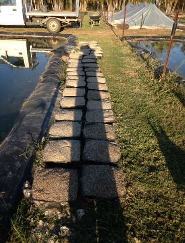 La imagen muestra un camino de piedra junto a una masa de agua, con una zona de césped a la derecha y un camión aparcado al fondo a la izquierda.