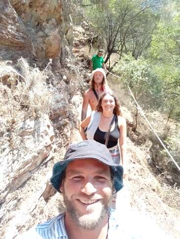 La imagen muestra a un grupo de cuatro personas subiendo por un sendero rocoso, rodeados de árboles y arbustos, bajo la luz del sol.