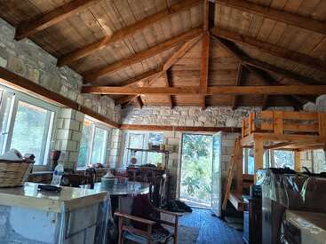 Essa aconchegante cabana de pedra tem tetos altos de madeira, janelas grandes, um beliche, uma mesa de jantar rústica, prateleiras abertas e uma convidativa porta aberta que revela a vegetação do lado de fora.