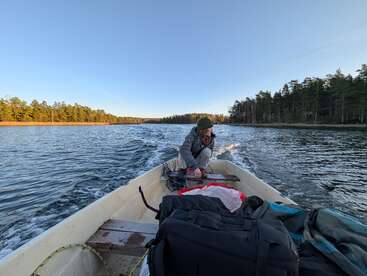 Una persona conduce una pequeña lancha motora por un lago tranquilo, rodeado de densos bosques y un cielo azul despejado, con aparejos y bolsas colocados en el interior de la embarcación.