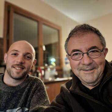 The image shows two men smiling at the camera, with one wearing a dark sweater and the other in a black jacket, in a room with a mirror and wooden door frame.