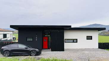 A modern, single-story house features black and white exterior walls, a bright red front door, flat roof, large windows, and a black electric car parked outside.