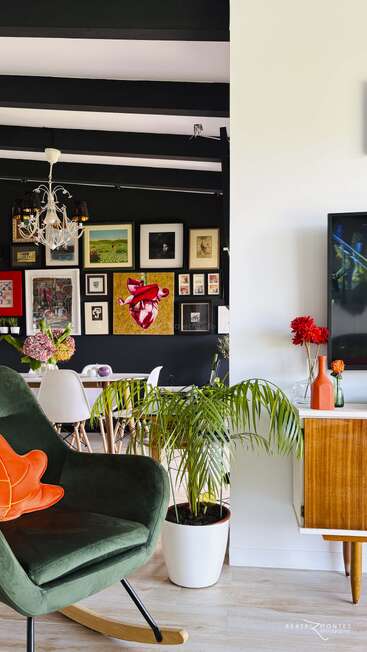 Eclectic living space with green rocking chair, orange pillow, indoor plants, wooden sideboard, TV, colorful wall art, and dining area with white chairs and chandelier.