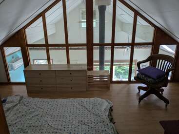 Dieses gemütliche Loft verfügt über eine Matratze auf dem Holzboden, eine Holzkommode, eine Kiste, einen gepolsterten Stuhl und große Fenster mit Blick auf den Raum darunter.