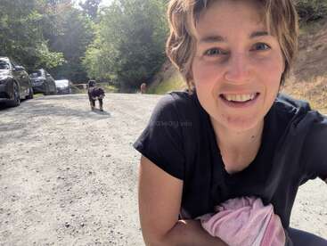 Eine lächelnde Frau macht auf einer sonnigen Schotterstraße ein Selfie und hält dabei ein rosa Tuch in der Hand. Im Hintergrund sind Autos und zwei Hunde zu sehen, umgeben von Bäumen.