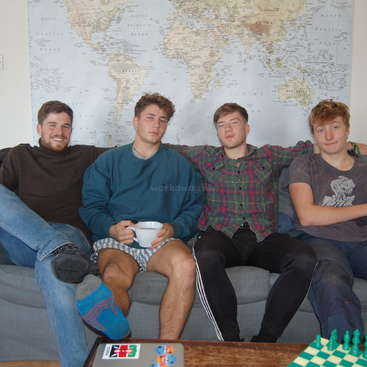 Das Bild zeigt vier junge Männer, die auf einer Couch vor einer Weltkarte sitzen, mit einem Schachbrett und einem Laptop auf einem Tisch im Vordergrund.