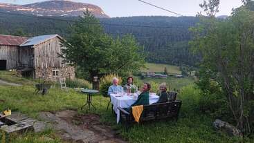 Vier Leute sitzen draußen an einem mit weißem Tischtuch gedeckten Tisch und genießen ihr Essen. Der Blick fällt auf ein üppig grünes Tal, eine alte Holzscheune und die Berge in der Ferne.