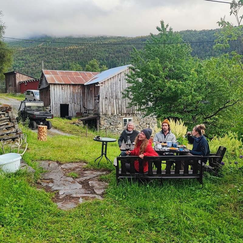 Vier Leute sitzen an einem Holztisch im Freien und genießen Essen und Getränke in einer üppig grünen, ländlichen Umgebung mit rustikalen Scheunen, Bäumen und nebelverhangenen Bergen.
