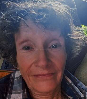 Ein Selfie einer älteren Person mit lockigem Haar, die ein kariertes Hemd trägt und sanft lächelt, in Nahaufnahme. Weiches natürliches Licht hebt ihr Gesicht und ihren freundlichen Ausdruck hervor.