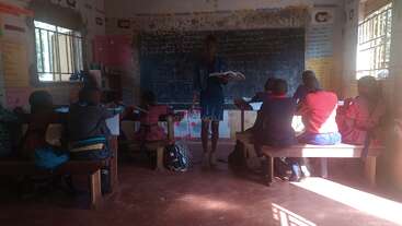 A teacher stands at the front of a classroom, reading from a book to students sitting on benches, engaging with their lessons in a sunlit room.