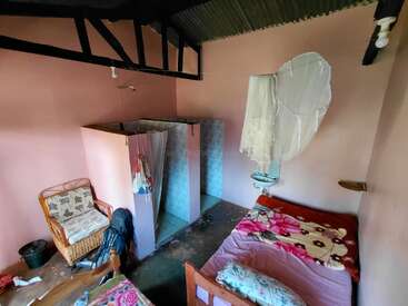 This image shows a small, simple room with pink walls, a single bed with floral blankets, a mosquito net, basic bathroom, wicker chair, and exposed beams.